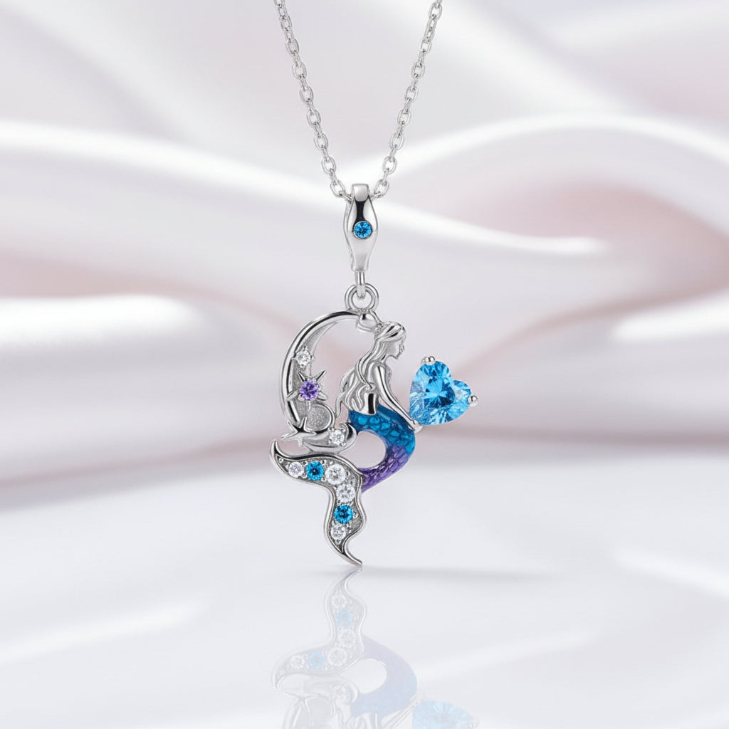 A silver pendant necklace featuring a mermaid design with a crescent moon, purple and blue enamel details, and sparkling blue and clear stones, shown on a soft white and pink satin background.