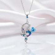A silver pendant necklace featuring a mermaid design with a crescent moon, purple and blue enamel details, and sparkling blue and clear stones, shown on a soft white and pink satin background.