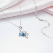 A silver-toned delicate necklace with a charm featuring a blue and purple bow and a small silver figure, displayed on a white surface with soft pink satin fabric in the background.