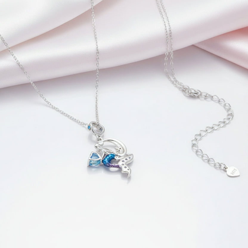 A silver-toned delicate necklace with a charm featuring a blue and purple bow and a small silver figure, displayed on a white surface with soft pink satin fabric in the background.