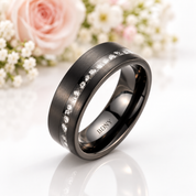 A dark gunmetal gray tungsten band ring with a channel-set row of clear round stones, featuring the engraving 'BDNY' on the inside, displayed on a reflective white surface with soft pink and white flowers in the background.