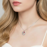 A close-up of a woman wearing a delicate silver necklace with a heart-shaped pendant featuring a small red gemstone accent.