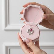 A silver pendant necklace with a small red heart is displayed inside an open pink jewelry box, held by two hands against a soft white paneled background.