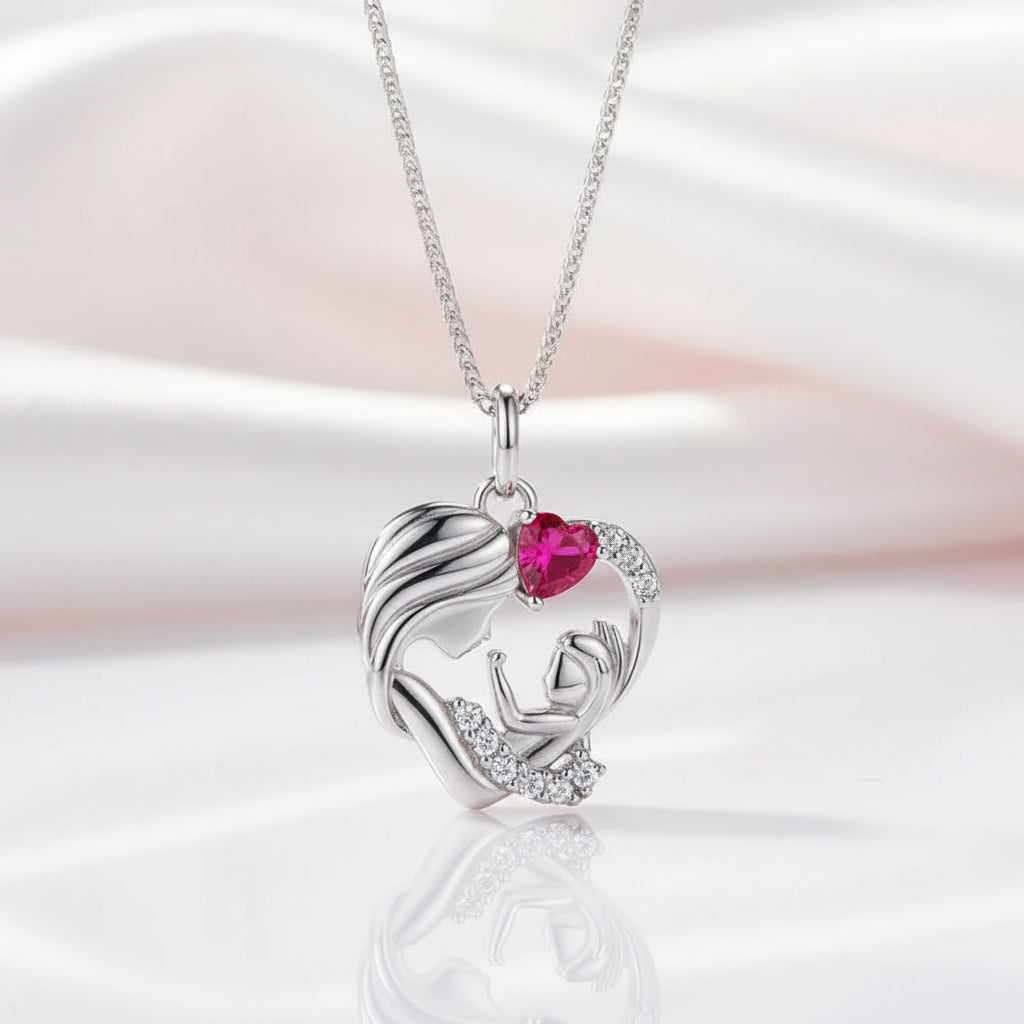 A silver pendant necklace featuring a mother and child design with a heart-shaped pink gemstone and small clear stones, displayed on a soft white and pink satin background.