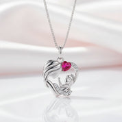 A silver pendant necklace featuring a mother and child design with a heart-shaped pink gemstone and small clear stones, displayed on a soft white and pink satin background.