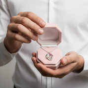 A silver horse pendant necklace with a pink gemstone is displayed inside an open light pink jewelry box held by a person wearing a white button-down shirt.
