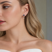 A close-up of a woman wearing a delicate cross-shaped dangle earring with dark blue stones and a small clear teardrop charm, shown against her ear and blonde hair.