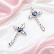 A pair of silver-toned dangle earrings with heart-shaped iridescent stones and small clear crystal accents, displayed on a glossy white surface with soft pink satin fabric.
