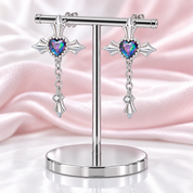 A pair of silver-toned gothic cross dangle earrings with heart-shaped iridescent gemstones, displayed on a silver stand against a pink satin background.