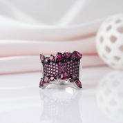 A silver ring featuring a wide band paved with deep pink gemstones and accented by larger, faceted pink stones, displayed on a reflective white surface with soft pink and white background elements.