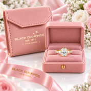 A gold ring with a large oval clear gemstone is displayed in an open pink velvet jewelry box, with a matching closed pink jewelry pouch and ribbon in the background. The boxes and ribbon are branded BLACK DIAMONDS NEW YORK.