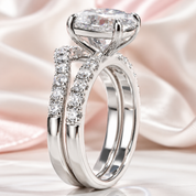 A silver-toned split-shank engagement ring with a large round brilliant-cut center stone and pavé-set accent stones, displayed on a soft pink satin fabric background.