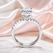 A silver-toned solitaire engagement ring with a large round brilliant-cut center stone and a pavé-set band of small clear stones, displayed on a soft pink satin fabric background.