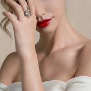 A close-up lifestyle shot of a model wearing a silver snake ring on her finger, with her hand resting near her face. She is wearing a white off-the-shoulder top against a soft beige background.