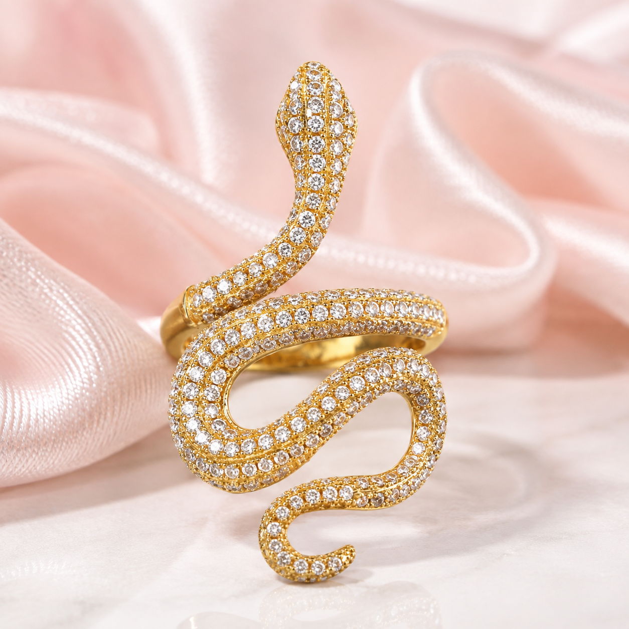 A gold snake ring paved with clear cubic zirconia stones, resting on a soft pink satin fabric background.