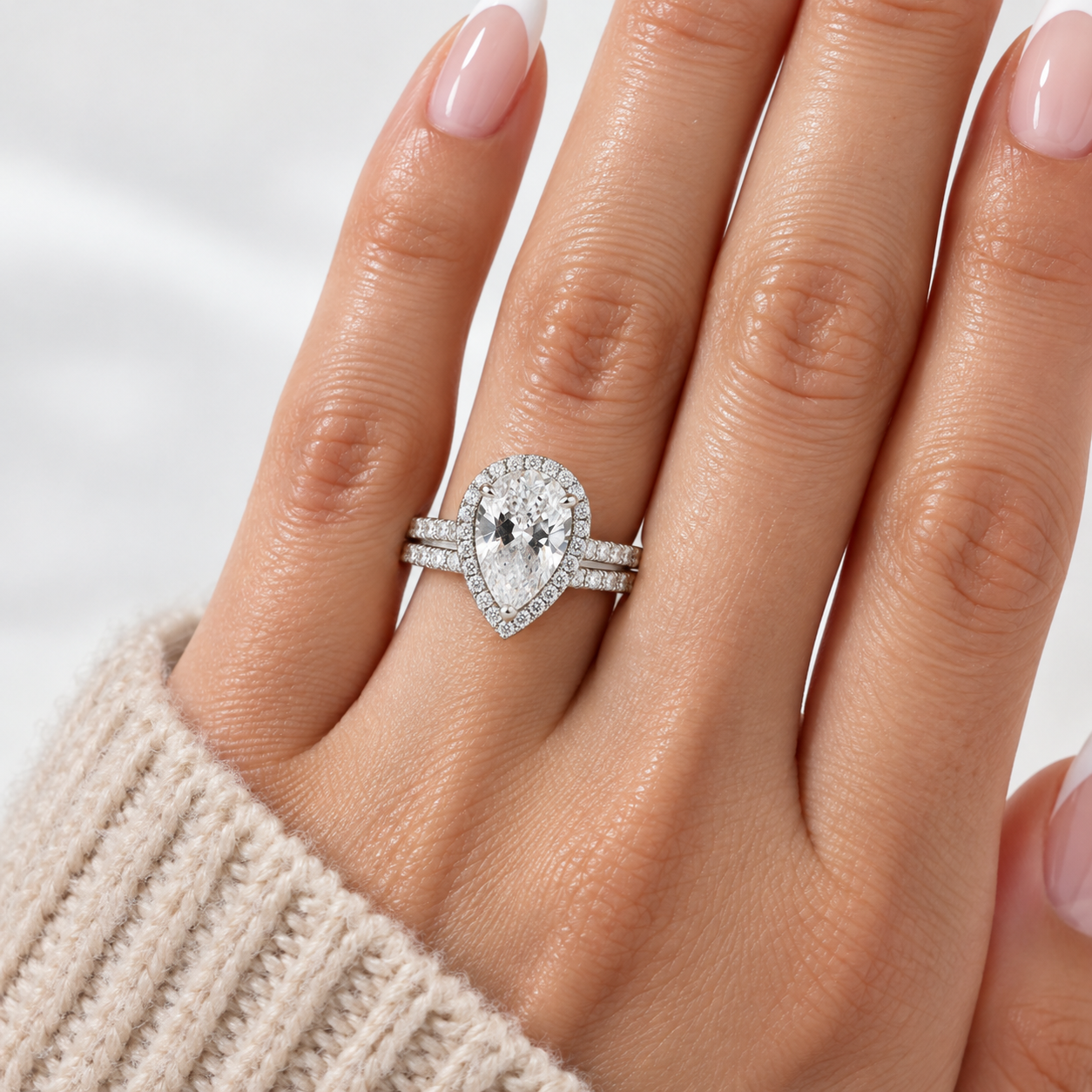 A silver-tone ring with a pear-cut center stone and a halo of small clear stones, shown worn on a hand with pale pink manicured nails and a beige knit sleeve visible.