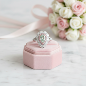 A pear-cut green gemstone ring with a halo of diamonds and pink accent stones, displayed on a pink velvet ring box with a bouquet of white and pink roses in the background.