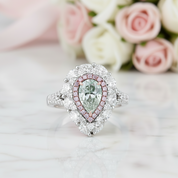 A pear-cut diamond halo ring in white metal with a pink diamond border, displayed on a light marble surface with soft pink and white roses in the background.