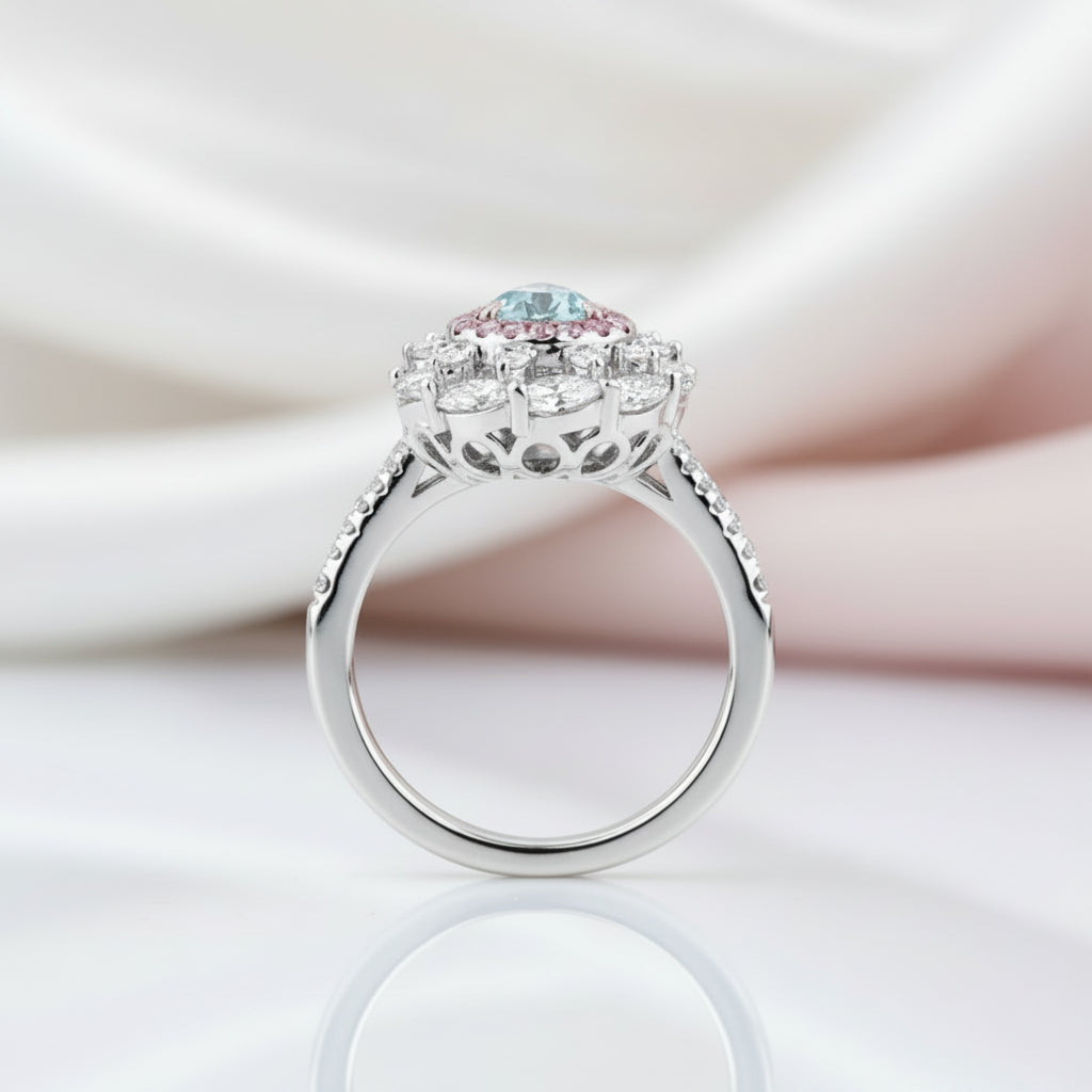A silver-toned halo ring featuring a central round light blue gemstone accented by small pink and clear stones, displayed on a reflective white surface with soft draped satin fabric in the background.