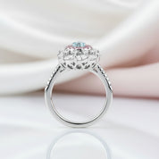 A silver-toned halo ring featuring a central round light blue gemstone accented by small pink and clear stones, displayed on a reflective white surface with soft draped satin fabric in the background.