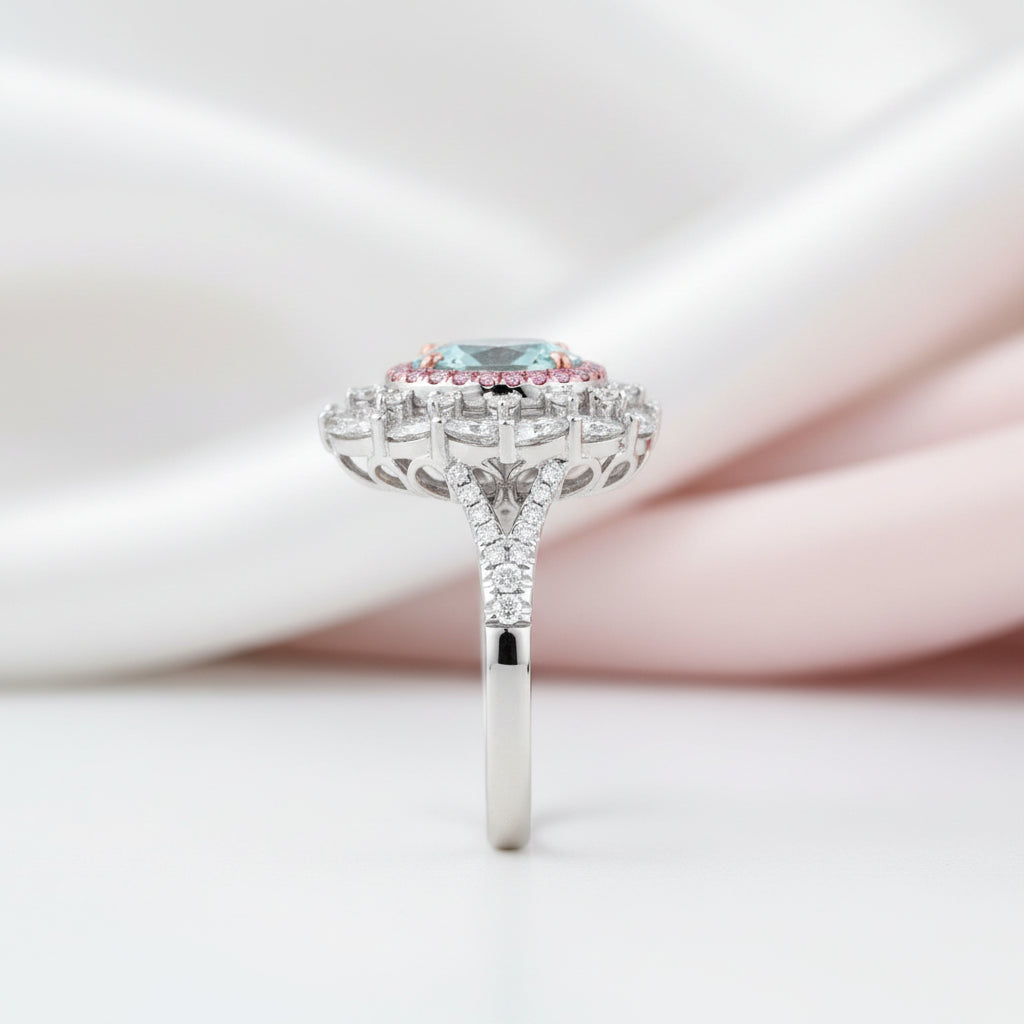 A silver-toned halo ring featuring a round light blue gemstone surrounded by small pink stones and clear accent stones, set against a soft white and pale pink draped fabric background.