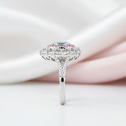 A silver-toned halo ring featuring a round light blue gemstone surrounded by small pink stones and clear accent stones, set against a soft white and pale pink draped fabric background.
