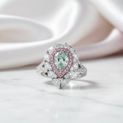 A pear-cut green gemstone ring with a halo of pink stones and white stones, set on a white band, resting on a white marble surface with a soft pink satin background.