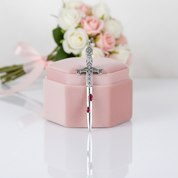 A silver-toned dagger-shaped ring with clear stones and small red gem accents, displayed upright on top of a pink jewelry box with blurred pink and white roses in the background.
