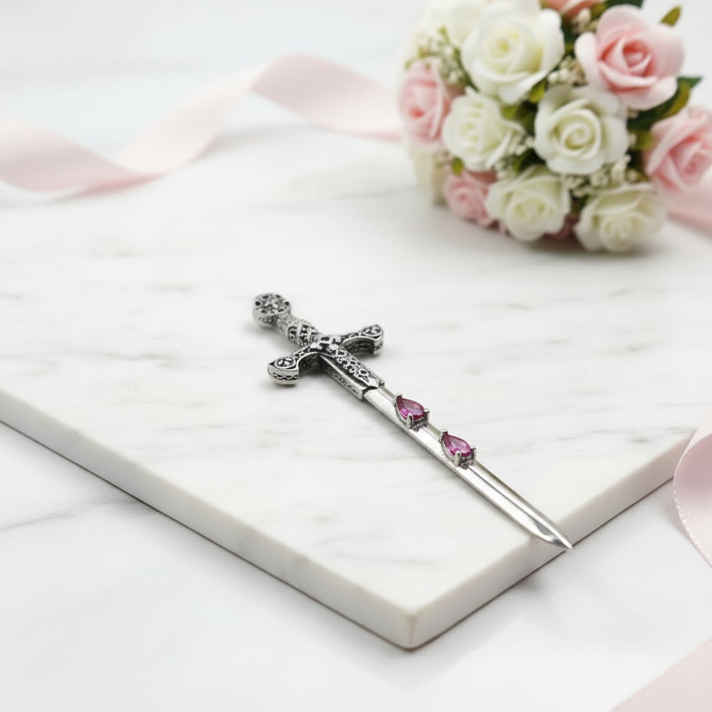 A silver-toned decorative letter opener styled like a miniature sword, featuring three small pink gemstones along the blade, resting on a white marble slab with pink roses in the background.
