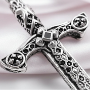 Close-up of an ornate silver-toned cross pendant with intricate filigree cutouts and a central square black inlay, photographed on a plain light background.
