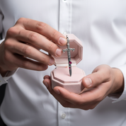 A close-up of a person in a white dress shirt holding an open light pink jewelry box and presenting a silver cross pendant with small pink gemstones.