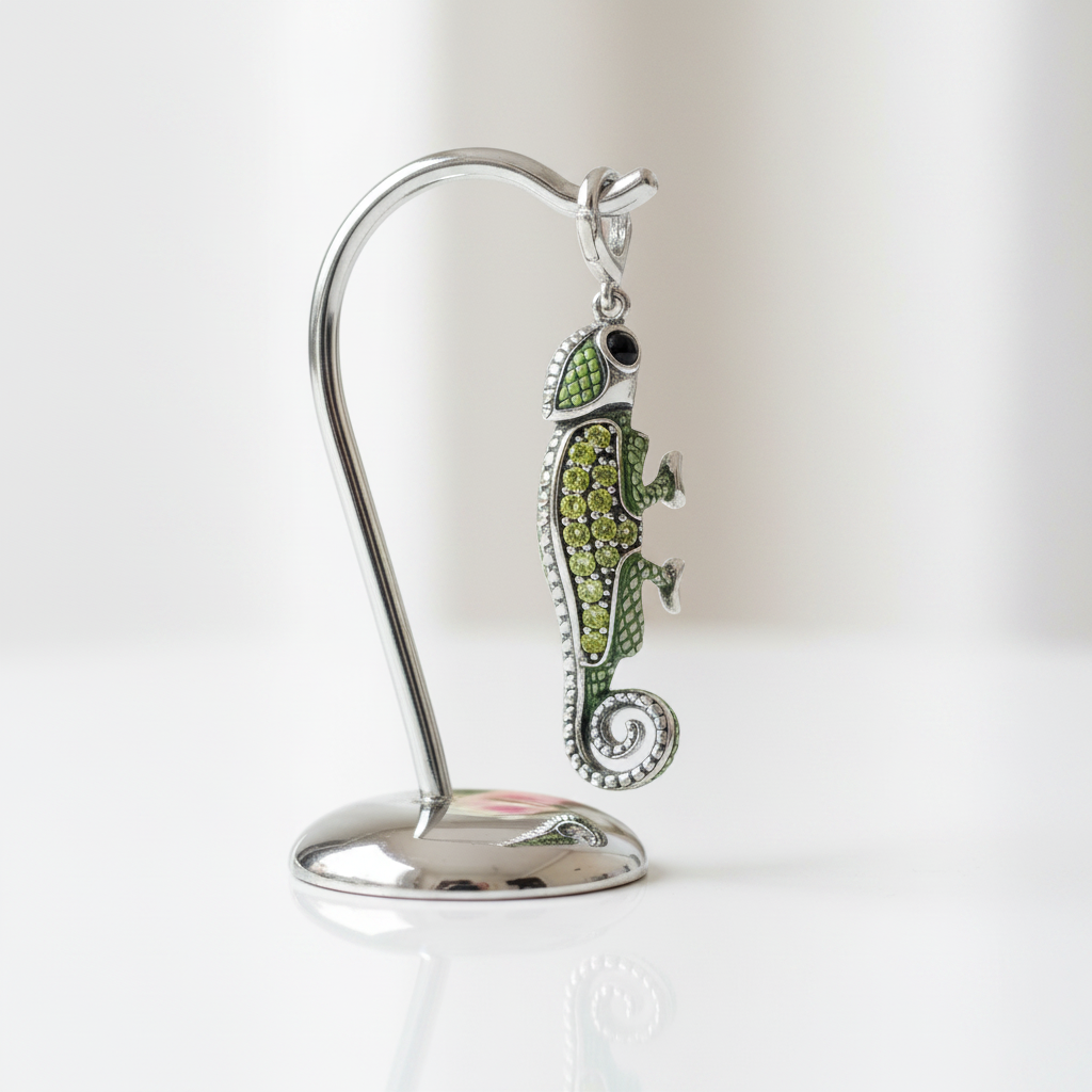 A silver-toned chameleon pendant with green enamel and rhinestone detailing, displayed on a curved silver stand against a soft white background.