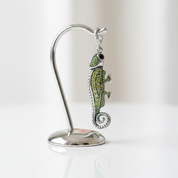 A silver-toned chameleon pendant with green enamel and rhinestone detailing, displayed on a curved silver stand against a soft white background.