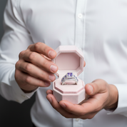 Person holding a pink hexagonal ring box with a ring inside, wearing a white shirt.