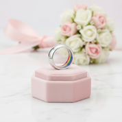 A polished silver band ring with a rainbow-colored interior sits on a small pink velvet ring box, with a blurred bouquet of white and pink roses in the background.