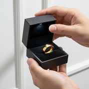 Gold ring with a rainbow design in an open black box held by two hands.