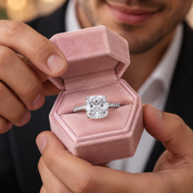A close-up of a man holding an open pink velvet ring box that displays a silver-toned ring with a large cushion-cut diamond. The man is wearing a black suit jacket and white collared shirt.