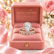 A rose gold teardrop-shaped diamond ring with a matching pavé band, presented in an open pink velvet Black Diamonds jewelry box with decorative pink roses and ribbons in the background.