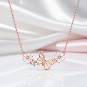 Rose gold necklace featuring a central butterfly pendant flanked by two flower motifs and a small clear crystal cluster, displayed on a soft white satin fabric background.