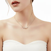 A close-up of a woman wearing a delicate rose gold necklace with a small floral cluster pendant, styled with a white strapless top against a clean white background.