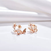 A pair of rose gold butterfly and flower drop earrings with a small clear crystal accent, displayed on a clean white textured surface.