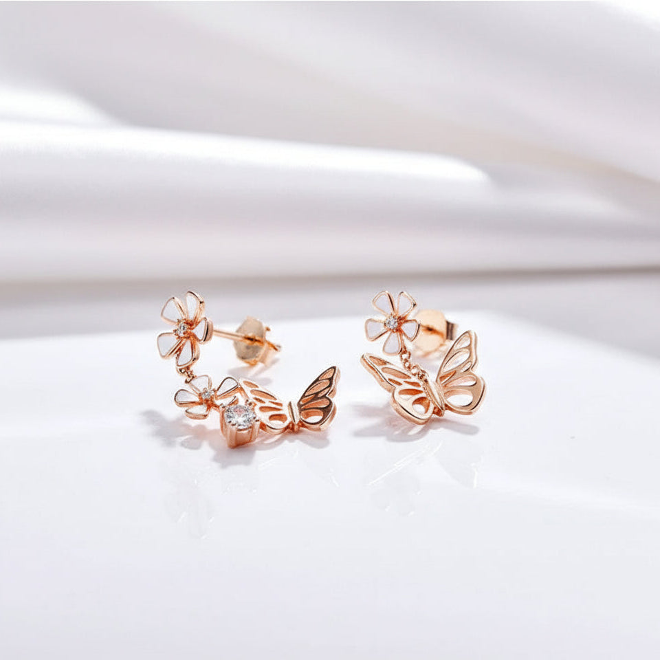 A pair of rose gold butterfly and flower drop earrings with a small clear crystal accent, displayed on a clean white textured surface.