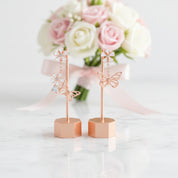Two rose gold butterfly and flower jewelry display stands on a white marble surface, with a blurred bouquet of pink and white roses in the background.