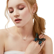 A close-up lifestyle shot of a woman with blonde hair tied back, wearing a delicate silver necklace and a silver ring adorned with a large, iridescent blue and green butterfly. She is wearing a white strapless top against a clean white background.