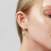 A close-up of a woman's ear wearing a silver dangle earring with a flower stud and a butterfly charm, set against a soft light gray background.