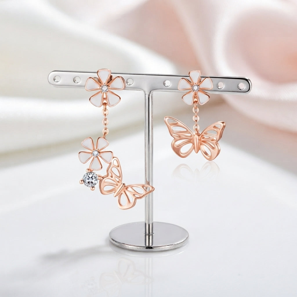 Rose gold butterfly and flower dangle earrings displayed on a silver T-bar earring stand against a soft white and pale pink draped fabric background.