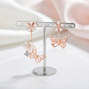 Rose gold butterfly and flower dangle earrings displayed on a silver T-bar earring stand against a soft white and pale pink draped fabric background.