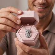 A close-up lifestyle shot of a man holding an open pink jewelry box that displays a silver pendant necklace with a cat head design set with clear gemstones and green accents.