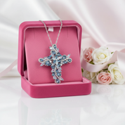 Silver cross necklace with blue stones in a pink jewelry box on a white background with flowers.