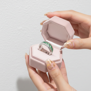 Ring with green gemstone in a pink jewelry box held by hands against a light gray background
